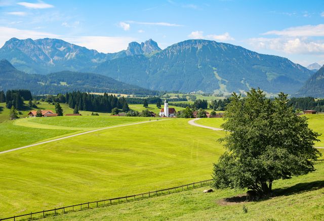 This image captures a serene countryside landscape with rolling green fields, a quaint church in the distance, and majestic mountains in the background. Ideal for travel brochures, nature-inspired content, or rural tourism promotions.