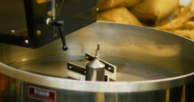 Close-up view of an industrial coffee roasting drum actively processing fresh beans. Perfect for use in food and beverage industry advertisements, coffee shop promotions, machinery and equipment catalogs, and educational materials about coffee production.