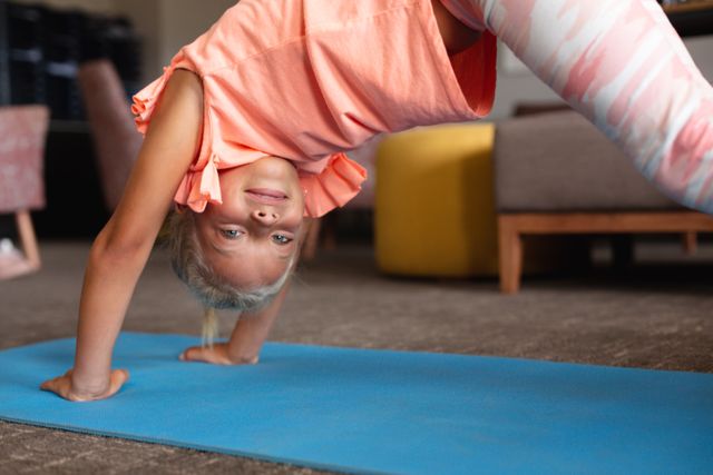 Young girl practicing yoga on a blue mat indoors. She is smiling and appears to be enjoying the activity. Ideal for use in educational materials, fitness programs for children, or promoting healthy lifestyles and physical activities for kids.