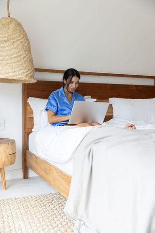 Woman in blue pajamas working on laptop on wooden bed in minimalist cozy bedroom