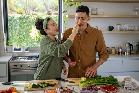 Couple preparing vegetables and laughing while sharing snack on bright modern kitchen island
