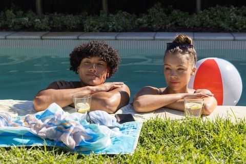 Young couple relaxing at backyard pool edge with lemonade and inflatable beach ball on sunny afterno