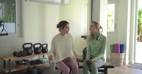 Female workout partners sitting on bench chatting and hydrating in bright home gym