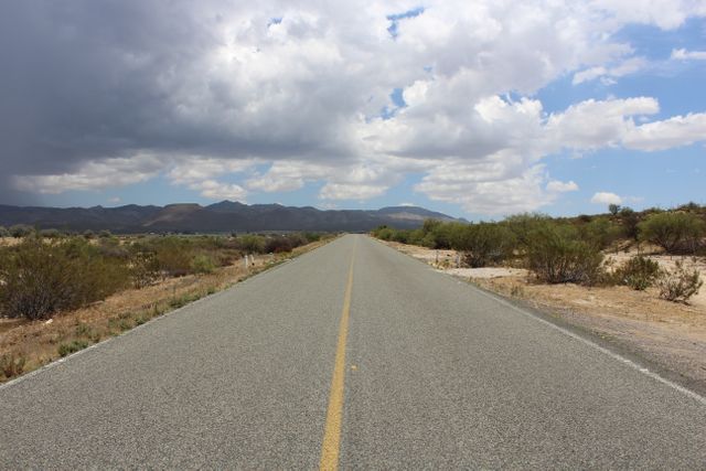 Solitary desert road leading towards distant mountains with dramatic clouds overhead, perfect for themes of travel, adventure, and solitude. Ideal for websites, blogs, or advertisements promoting road trips, outdoor experiences, or rural exploration.