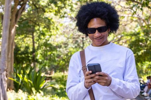 Adult man using smartphone in sunny park wearing sunglasses and crossbody bag strap