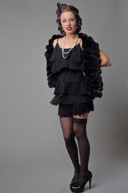 Authentic portrayal of a confident woman in 1920s flapper style outfit with layered black dress, fur shawl, pearls, and high heels. Useful for themes involving vintage fashion, roaring twenties, costume parties, or historical fashion retrospectives.