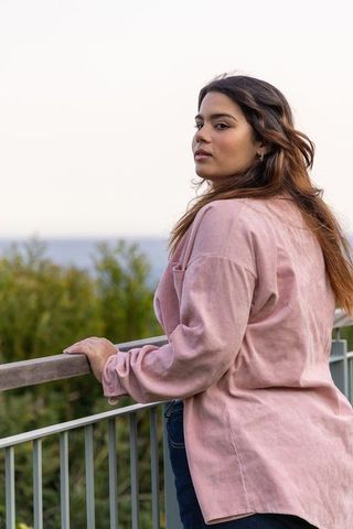 Young woman standing at railing overlooking sea in pink overshirt, glancing back