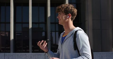 Young man walking in city listening with wireless earbuds while holding smartphone
