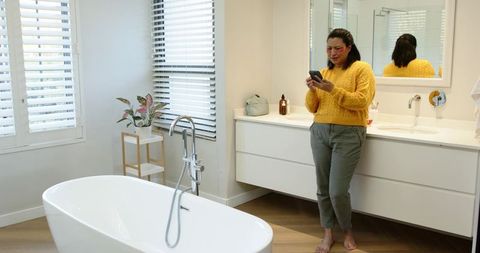 Woman Using Smartphone in Minimalist Bathroom, Next to Bathtub