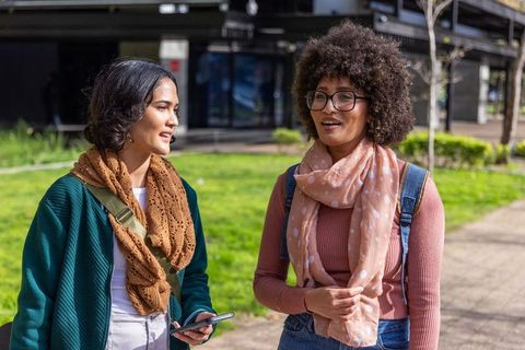 Two diverse female students walking and chatting on university campus holding smartphone
