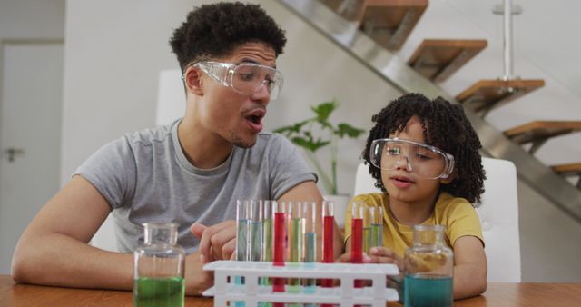 Father and young son conducting a science experiment at home. Both wearing protective goggles and looking at colorful liquids in test tubes. Ideal for educational content, promoting STEM activities, family bonding and learning resources.