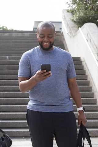 Smiling african american man checking smartphone on urban steps with bag and smartwatch