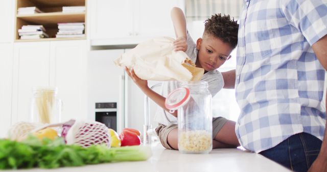 Boy actively participating in kitchen chores by pouring cereal into jar with parent's assistance. Useful for concepts of family bonding, parenting tips, child development, and home cooking.