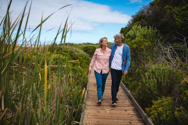 Senior couple walking arm in arm on a wooden bridge surrounded by lush greenery. Ideal for use in lifestyle, retirement, and health-related content. Perfect for illustrating themes of love, togetherness, and active living in senior years.