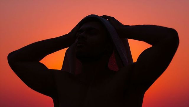 Shirtless man draping towel over head at sunset silhouette vibrant orange sky dusk