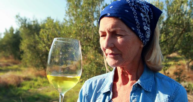 Elderly woman holding a glass of white wine while standing in a lush vineyard. She is savoring the moment, dressed casually in a blue bandana and denim shirt, with a content expression. Perfect for content related to wine culture, senior activities, relaxation, outdoor lifestyles, vineyards, and wine tasting experiences.