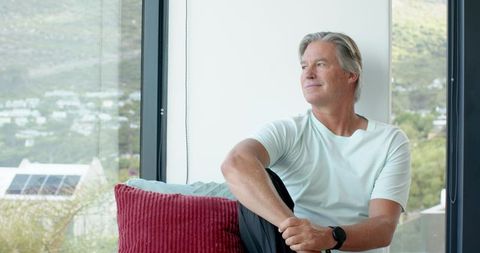 Senior man relaxing on window seat gazing at hillside solar panels in modern daylight home