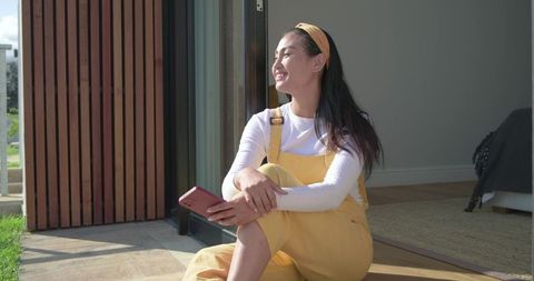 Mid-adult asian woman in yellow overalls smiling in sunlight while sitting at doorway threshold