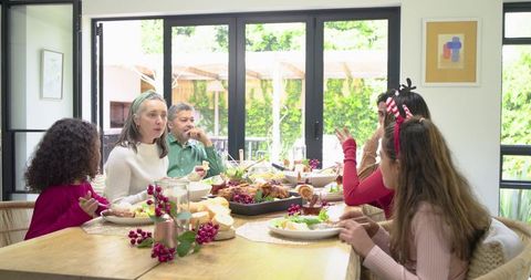 Multi-generation family sharing sunlit holiday meal around wooden table, passing roasted platter
