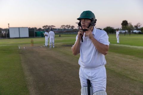 Batsman fastening helmet chin strap on cricket pitch at dusk