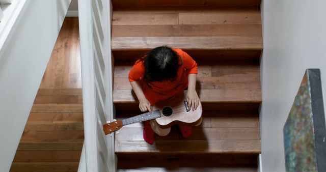 Young child sitting on wooden stairs playing a ukulele, viewed from above. Child is wearing casual clothing in a well-lit home. Perfect for themes of childhood, music practice, home interiors, and leisure activities.