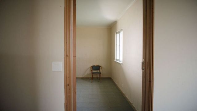 Minimalist empty room with lone blue chair by window viewed through open doorway