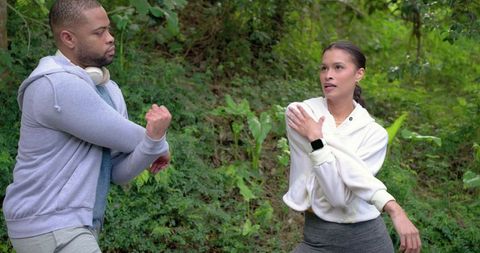 Diverse couple stretching arms across chest on wooded trail in hoodies and activewear