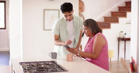 Young man helping senior woman use smartphone in kitchen environment