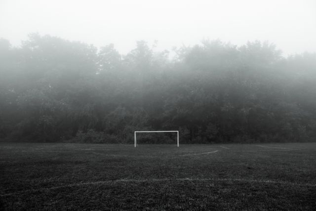 This image depicts a foggy soccer field in the early morning, with an empty goalpost set against a backdrop of trees and mist. Suitable for illustrating concepts related to sports, tranquility, atmospheric scenes, and natural landscapes. Ideal for use in blog posts about early morning activities, sports, mindfulness, and nature.