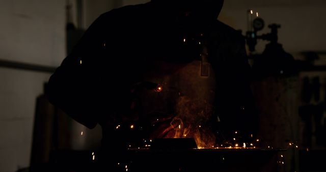 Silhouette of a metal worker welding, sending sparks flying, in a dimly lit industrial workshop. This image, rich with contrast and shadow, captures the essence of intense manual labor and skilled craftsmanship. Ideal for use in industrial, engineering, DIY, and manual labor-related projects, showcasing the gritty, hands-on work of a dedicated welder or blacksmith.