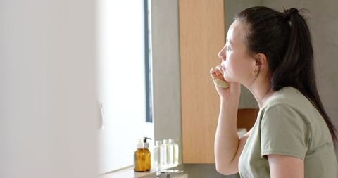 Asian woman applying face cream with puff at modern bathroom sink, morning skincare