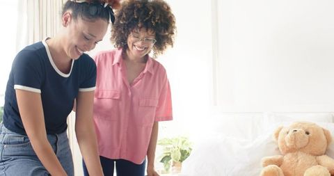 Friends arranging clothes on sunlit bedroom bed with teddy bear and plant