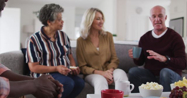 Group of seniors and middle-aged friends gathering in cozy home environment. People are casually chatting while holding mugs and enjoying snacks at coffee table. Ideal for depicting social interaction, friendship, healthy relationships, and comfortable home setting.