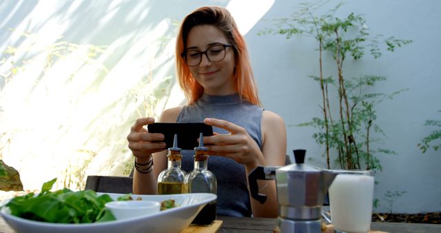 Young woman enjoying coffee while using smartphone outside. Smiling and wearing glasses seated at an outdoor table with fresh salad and condiments. Ideal for leisure, outdoor dining, technology or modern lifestyle concepts.