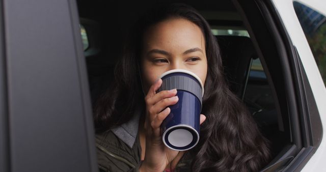 Asian woman with lowered face mask looking out of the window and drinking coffee sitting in the car. coronavirus covid-19 pandemic concept
