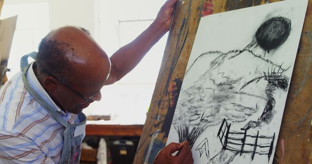 An African American senior man is focused on creating a charcoal drawing on paper, with copy space. His artistic expression is captured in a studio environment, highlighting his passion for visual arts.