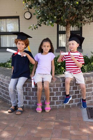 Children sitting on brick planter wearing mortarboards and holding red diplomas
