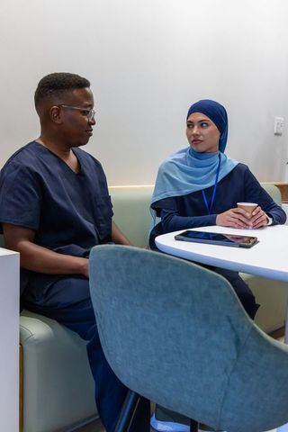 Healthcare team sitting in lounge wearing navy scrubs and hijab using tablet and coffee