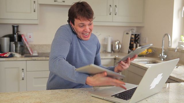 Man enthusiastically handling a busy work situation involving using a laptop, a mobile phone against his ear, and two tablets in a kitchen setting, illustrating a hectic multitasking moment. Useful for concepts related to work-life balance, technology impact on daily tasks, and showcasing the chaos that comes with digital communication. Perfect for blogs, presentations, or articles concerning technology usage, remote work challenges, and efficiency methods.