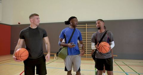 Walking diverse male athletes carrying basketballs and gear in indoor gym showing teamwork
