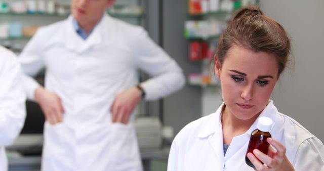 A Caucasian female pharmacist examines a medication bottle intently, with copy space. In the background, a male pharmacist, also Caucasian, is blurred, suggesting a busy pharmacy environment.