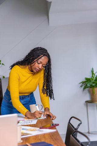 African american woman leaning over wooden table writing paperwork at home workspace with laptop