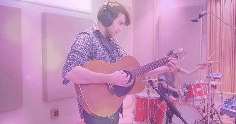 Young musician playing acoustic guitar in recording studio with drummer and microphones