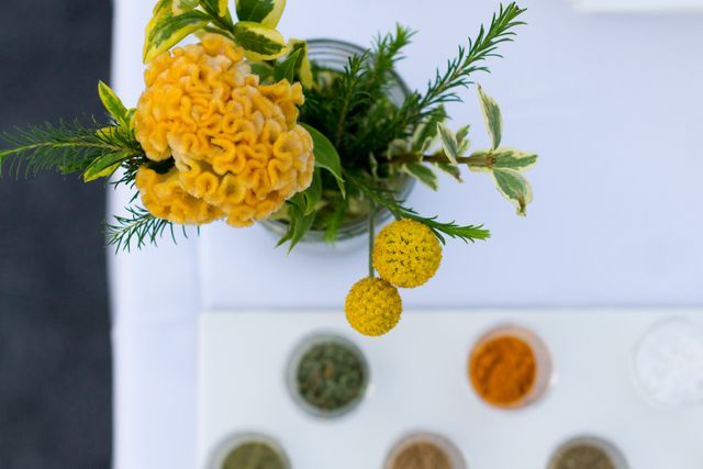 Aerial view of a vibrant yellow floral arrangement placed alongside a variety of dried herbs and spices on a crisp, white surface. Ideal image for promoting gardening services, culinary blogs or home decor ideas. This bright and refreshing setting can be utilized to highlight the beauty of natural ingredients in food and wellness content.