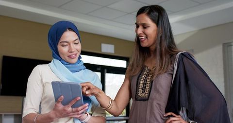Two indian women collaborating on tablet, one wearing hijab and one in embroidered tunic