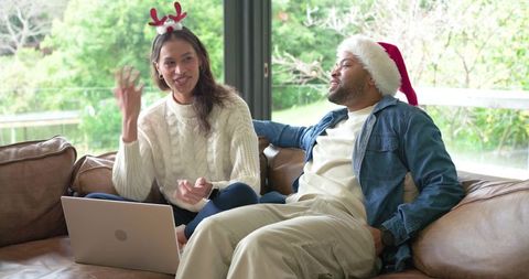 Diverse couple relaxing on sofa with laptop, santa hat and reindeer headband for holidays