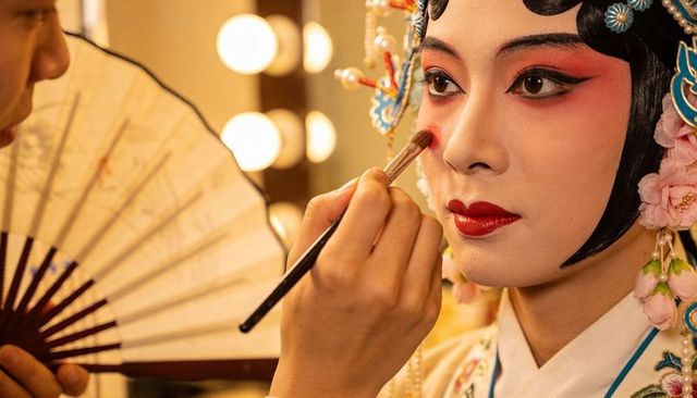 Peking opera performer receiving stage makeup backstage, traditional headdress and rouge
