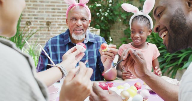 Multi-generational family members decorating Easter eggs together outside. Adults and children wearing bunny ears, sharing smiles and laughter. Perfect for use in themes such as Easter, family bonding, holiday decorations, and spring celebrations.