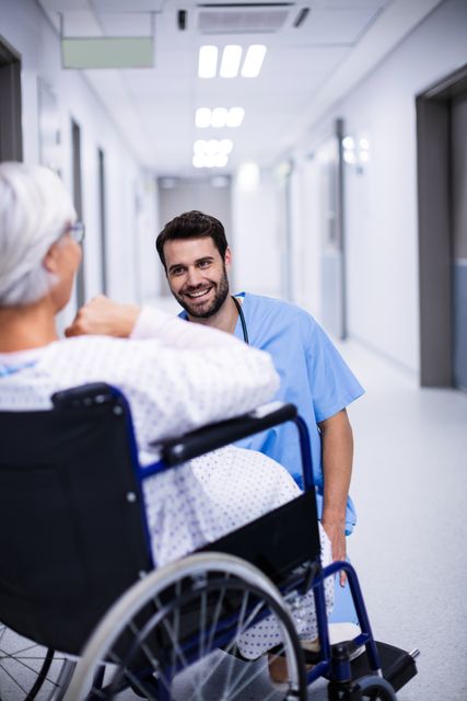 Male doctor kneeling and interacting with senior patient in wheelchair in hospital corridor. Ideal for use in healthcare, medical, and patient care contexts, highlighting compassionate care and professional medical services.