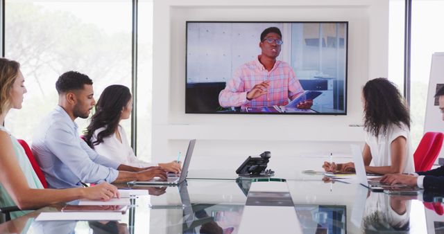 A diverse business team sitting at a conference table in a modern meeting room attending a virtual conference. They are paying attention to a colleague appearing on a screen, holding a tablet and presenting. Each person has a laptop in front of them, indicating an engaged and tech-savvy workplace. This image can be used for promoting remote collaboration tools, online meetings, corporate trainings, or any business communication aids.